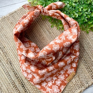May include: A rust-colored bandana with a white floral pattern. The bandana is tied in a knot and has a brown leather tag with the text "Happy & Free" on it.