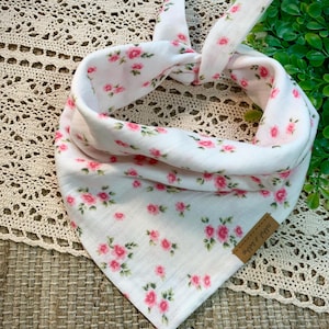 May include: White dog bandana with a pink floral pattern. The bandana is tied at the top, with a brown tag that reads "nibbit & sahha". The background is a woven mat and lace.