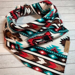 May include: A white, red, brown, black and turquoise bandana with a geometric pattern. The bandana has a small brown tag with the text "Wild & Free" on it.