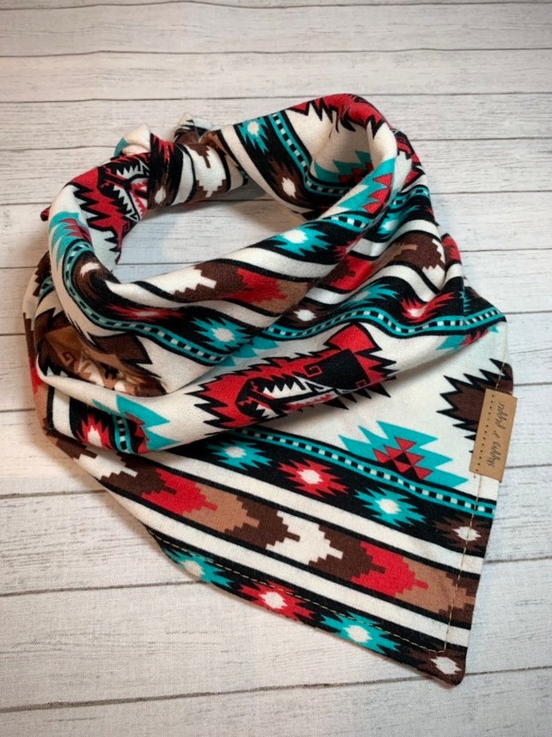 May include: A white, red, brown, black and turquoise bandana with a geometric pattern. The bandana has a small brown tag with the text "Wild & Free" on it.