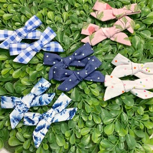 Könnte beinhalten: Eine Auswahl an Stoff-Haarschleifen in verschiedenen Mustern und Farben. Die Schleifen umfassen blaues Vichy-Karo, rosa Blumenmuster, Marineblau mit weißen Punkten, Weiß mit bunten Konfetti und blaue Blumenmuster. Die Schleifen sind auf einem Bett aus grünem Laub angeordnet.