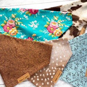 May include: Five different dog bandanas with various patterns. The bandanas are made of fabric and have a triangular shape. The patterns include floral, paisley, cow print, polka dots, and a geometric design. The bandanas are all light blue, brown, or white.