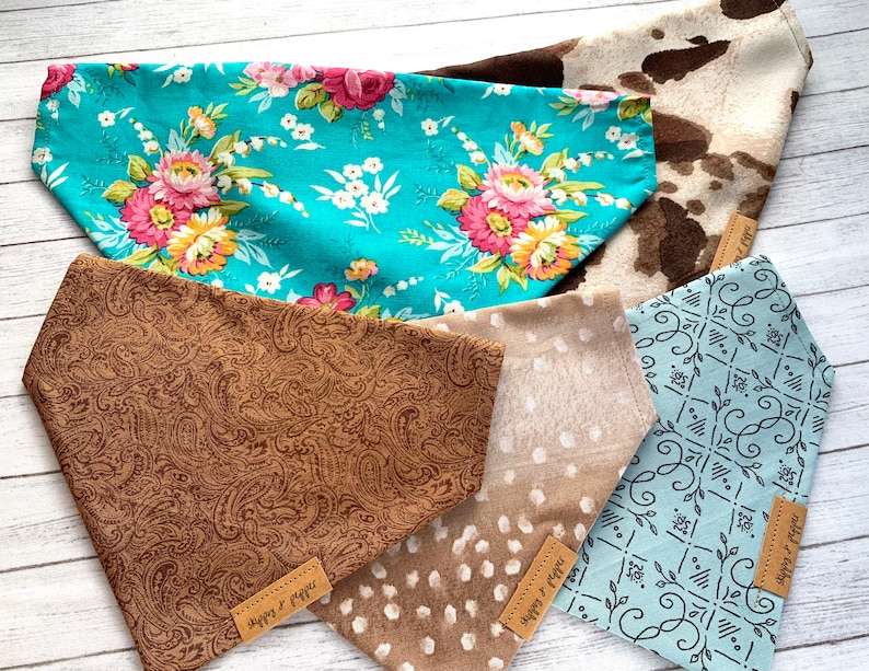 May include: Five different dog bandanas with various patterns. The bandanas are made of fabric and have a triangular shape. The patterns include floral, paisley, cow print, polka dots, and a geometric design. The bandanas are all light blue, brown, or white.