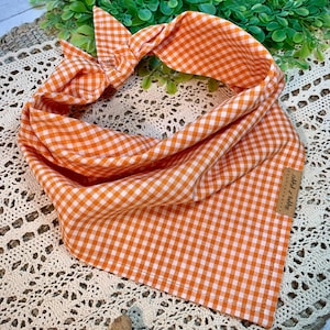 May include: Orange and white gingham dog bandana with a "Happy Tails" label attached. The bandana is tied in a knot and is lying on a crocheted surface.