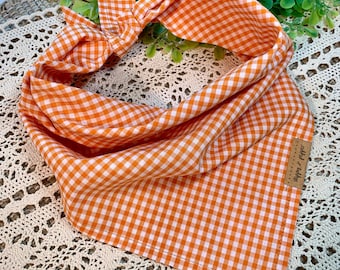 May include: Orange and white gingham dog bandana with a "Happy Tails" label attached. The bandana is tied in a knot and is lying on a crocheted surface.