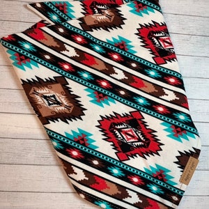 May include: A white bandana with a geometric pattern in shades of red, brown, turquoise, and black. The bandana is folded in a triangular shape and has a small brown tag with the text "Simply Southern" on it.