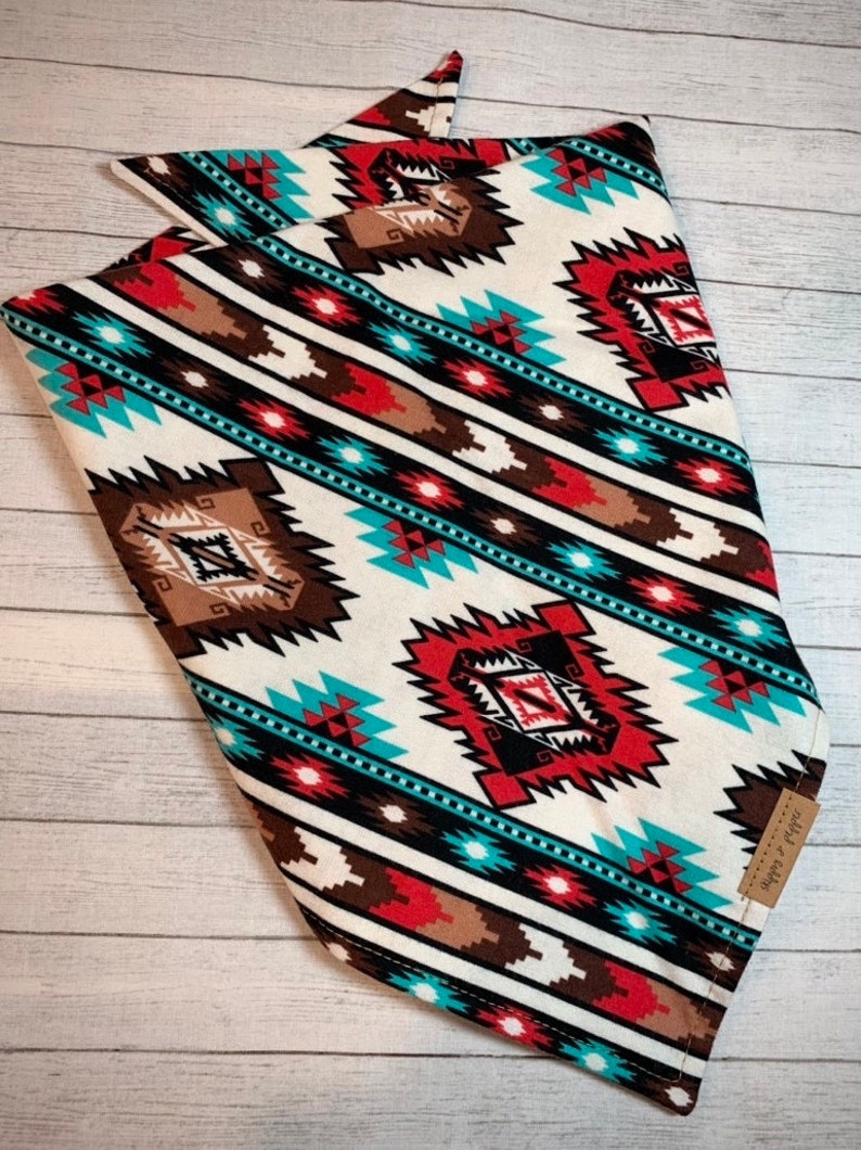 May include: A white bandana with a geometric pattern in shades of red, brown, turquoise, and black. The bandana is folded in a triangular shape and has a small brown tag with the text "Simply Southern" on it.