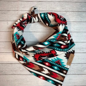 May include: A white bandana with a geometric pattern in shades of red, brown, turquoise, and black. The bandana has a brown leather tag with the text "shop by style" on it.