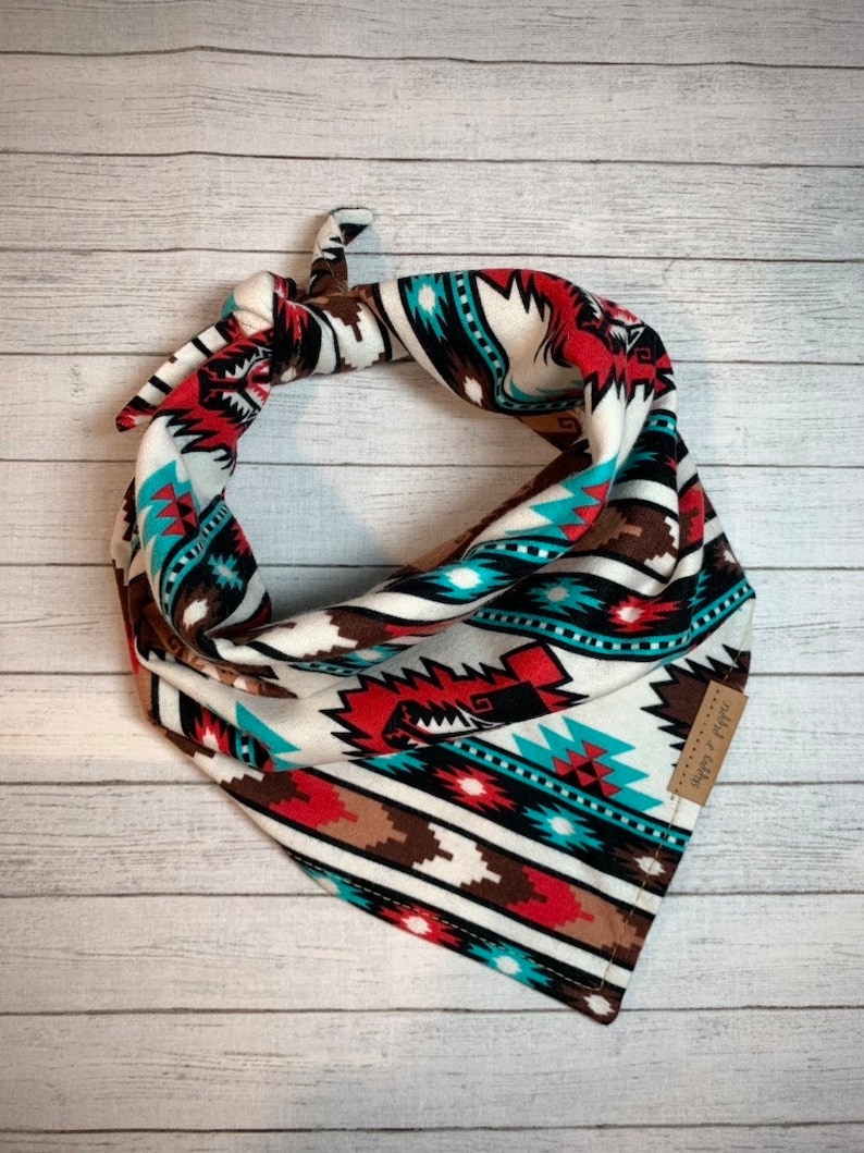 May include: A white bandana with a geometric pattern in shades of red, brown, turquoise, and black. The bandana has a brown leather tag with the text "shop by style" on it.