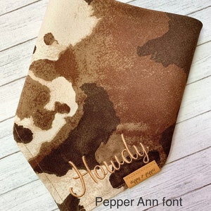 May include: A brown and tan bandana with a white watercolor-style pattern. The bandana is embroidered with the word "Howdy" in a tan thread. The bandana has a brown leather tag with the text "Stitches & Pepper" in white thread.