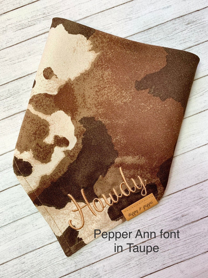 May include: A brown and tan bandana with a white watercolor-style pattern. The bandana is embroidered with the word "Howdy" in a tan thread. The bandana has a brown leather tag with the text "Stitches & Pepper" in white thread.