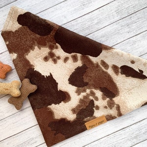 May include: Brown and white cow print dog bandana with a brown leather tag that says "Happy & Waggs".