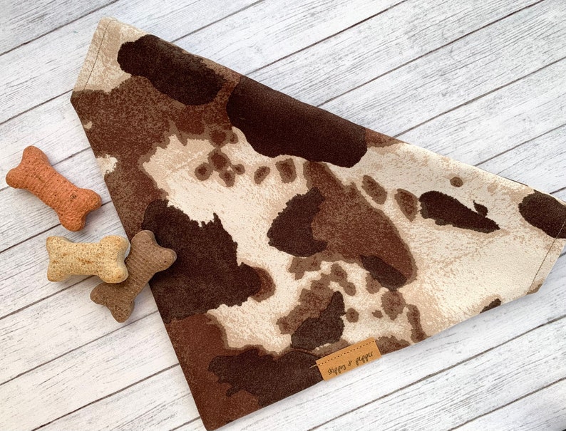 May include: Brown and white cow print dog bandana with a brown leather tag that says "Happy & Waggs".