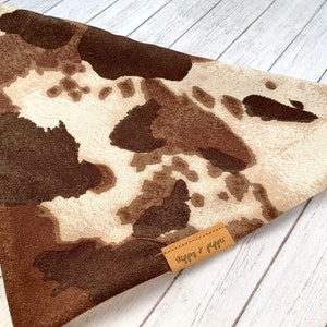 May include: A brown and white cow print bandana with a brown leather tag that says "Snaggs & Raggs".