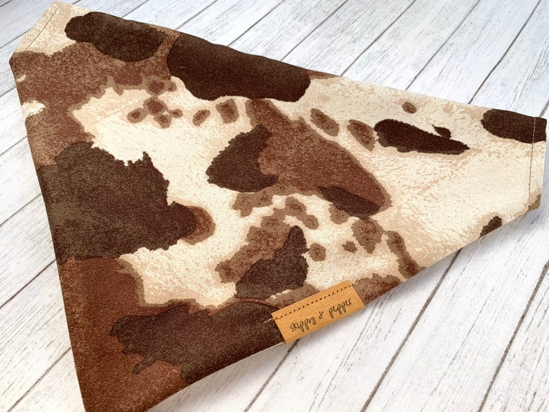 May include: A brown and white cow print bandana with a brown leather tag that says "Snaggs & Raggs".