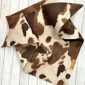 May include: A brown and white bandana with a cow print pattern. The bandana is folded into a triangle shape and has a small brown leather tag with the text "Happy Trails" on it.
