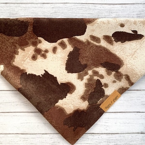 May include: A brown and white cow print bandana with a brown leather tag that says "Happy & Pawsitive".