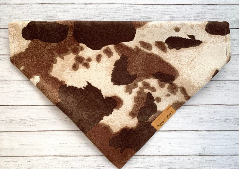 May include: A brown and white cow print bandana with a brown leather tag that says "Happy & Pawsitive".