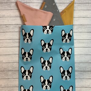 May include: A blue mailing bag with a repeating pattern of black and white French bulldog faces. The bag is filled with three fabric triangles, one pink, one grey with white arrows, and one yellow.