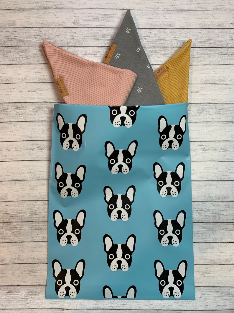 May include: A blue mailing bag with a repeating pattern of black and white French bulldog faces. The bag is filled with three fabric triangles, one pink, one grey with white arrows, and one yellow.