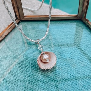 May include: A silver chain necklace with a pink seashell pendant. The seashell holds a pearl and a small crystal.