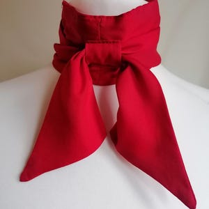 May include: A bright red neck scarf, knotted in a bow. The scarf is made of a smooth material and is shown on a white mannequin. The scarf's design includes a broad band that encircles the neck, with long, flowing ends.