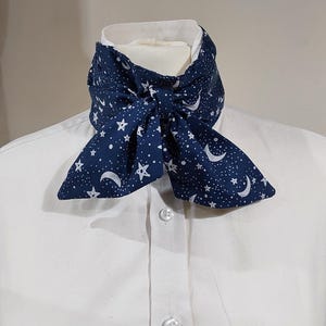 May include: A navy blue ascot featuring a white celestial design of stars and crescent moons, knotted in a bow. The ascot is worn over a white collared shirt. The fabric appears soft and lightweight, creating a unique and stylish look.