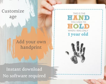 May include: A white card with the text "THIS IS THE HAND YOU USED TO HOLD WHEN I WAS ONLY 1 year old" in blue, orange, and green. A black handprint is below the text. The card is held by a person.