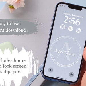 May include: A blue smartphone with a white background displaying a lock screen with the time 2:56, the date Monday, December 4, and the word "amelia" in a circle. The phone is being held by a hand.