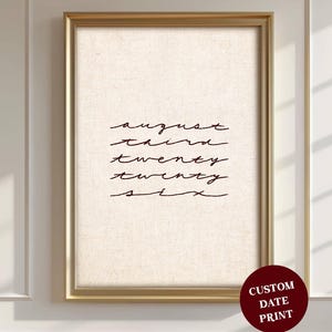 May include: A framed print featuring the date "august third twenty twenty six" in a handwritten font. The print is set within a gold frame, with a beige backdrop. A red circle with "CUSTOM DATE PRINT" is in the bottom right corner.