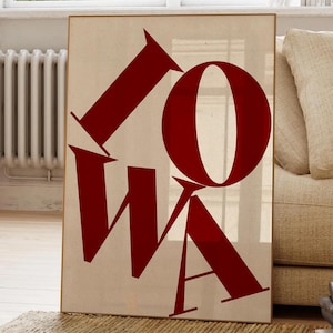 Puede incluir: Un póster enmarcado con la palabra "IOWA" en letras rojas y audaces sobre un fondo beige. Las letras están estilizadas y se superponen, creando un diseño gráfico moderno. El póster se exhibe en interiores, apoyado contra una pared.