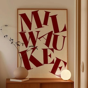May include: A poster with the word "MILWAUKEE" in large, red letters. It hangs above a wooden cabinet with a beige vase, books, and a spherical lamp. A sprig of greenery is in the vase. The room has a neutral colour palette.