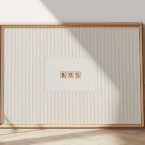 May include: A framed artwork featuring the letters "B.O.S." arranged on a cream-colored background with vertical stripes. The frame is a light wood color, and the artwork is displayed against a white wall with sunlight casting shadows.
