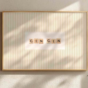 Puede incluir: Obra de arte enmarcada con la palabra "CINCIN" escrita con fichas de Scrabble. La obra está sobre un fondo a rayas en color beige claro, enmarcada en un marco dorado. La obra está colgada en una pared de color claro.