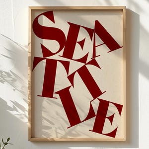 May include: A framed art print featuring the word "SEATTLE" in large, bold, red letters against a cream-colored background. The letters are arranged in a dynamic, overlapping design within a natural wood frame.