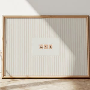 May include: A framed print displaying the letters "C, H, I" in Scrabble tiles. The print has a striped background and a light wood frame. The artwork is displayed on a wall with a wooden floor.