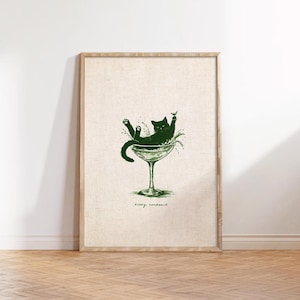 May include: A framed print featuring a green illustration of a cat lounging in a martini glass. The cat is surrounded by splashes, with the words "Kitty Cocktail" written below in a script font. The artwork is set against a neutral background.