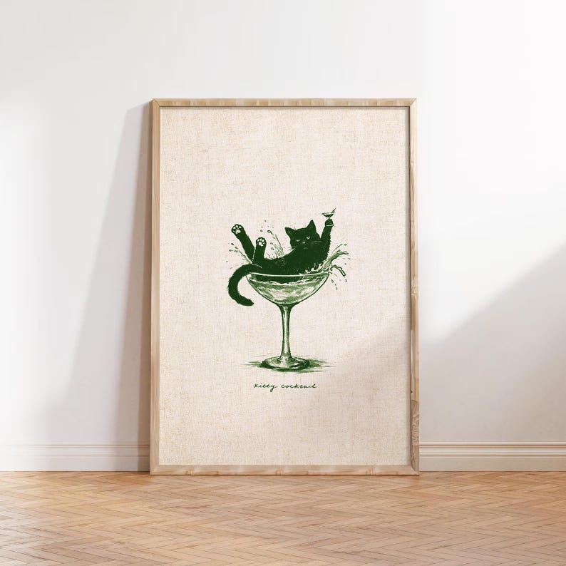 May include: A framed print featuring a green illustration of a cat lounging in a martini glass. The cat is surrounded by splashes, with the words "Kitty Cocktail" written below in a script font. The artwork is set against a neutral background.