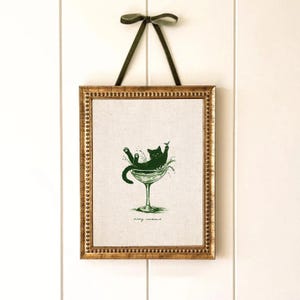May include: Framed art print featuring a green illustration of a black cat lounging in a martini glass. The print is in a gold-colored frame and is suspended by a green ribbon. The text "Kitty Cocktail" is at the bottom of the print.