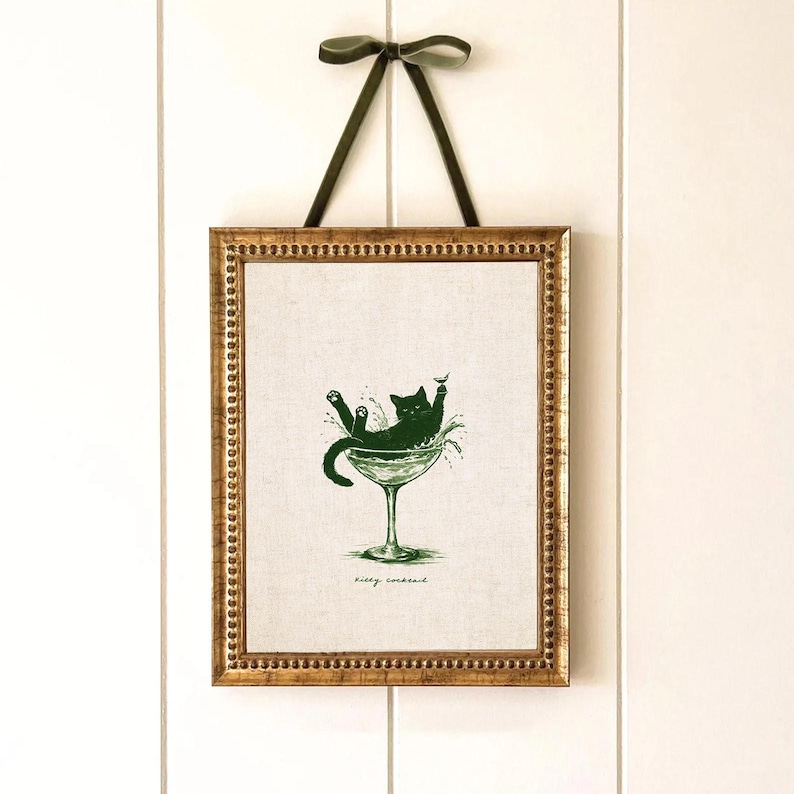 May include: Framed art print featuring a green illustration of a black cat lounging in a martini glass. The print is in a gold-colored frame and is suspended by a green ribbon. The text "Kitty Cocktail" is at the bottom of the print.