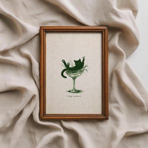 May include: A framed print featuring a green illustration of a cat lounging in a martini glass. The cat is black, and the glass is green. The text "kitty cocktail" is at the bottom of the print. The frame is made of brown wood.