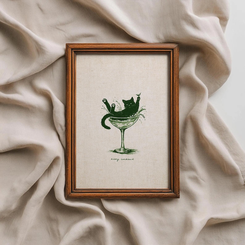 May include: A framed print featuring a green illustration of a cat lounging in a martini glass. The cat is black, and the glass is green. The text "kitty cocktail" is at the bottom of the print. The frame is made of brown wood.