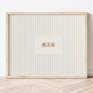Könnte beinhalten: Gerahmtes Kunstwerk mit dem Namen "MIA", geschrieben mit Scrabble-Steinen. Das Kunstwerk hat einen gestreiften Hintergrund in hellem Beige und ist in einem hellen Holzrahmen eingefasst. Das Design ist minimalistisch und dekorativ.