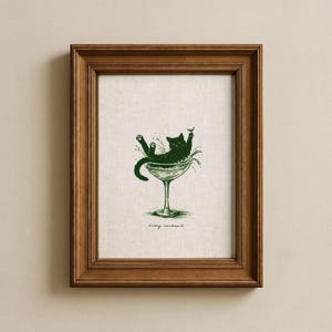 May include: A framed print featuring a green illustration of a black cat lounging in a martini glass. The cat is surrounded by splashes, with the words "kitty cocktail" below the glass. The frame is made of brown wood.