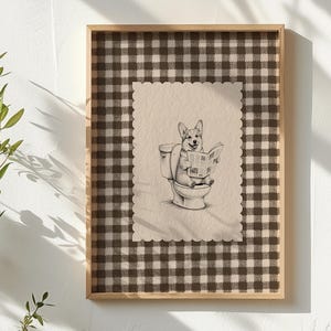 May include: A framed wall art print featuring a light brown and white illustration of a Corgi sitting on a toilet while reading a newspaper. The central image is on a scalloped-edge paper background, set against a dark brown and white gingham pattern.