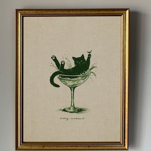 May include: Framed art print featuring a green illustration of a black cat lounging in a martini glass, with the words "Kitty Cocktail" below. The print is set against a neutral background and framed in a gold-colored frame.