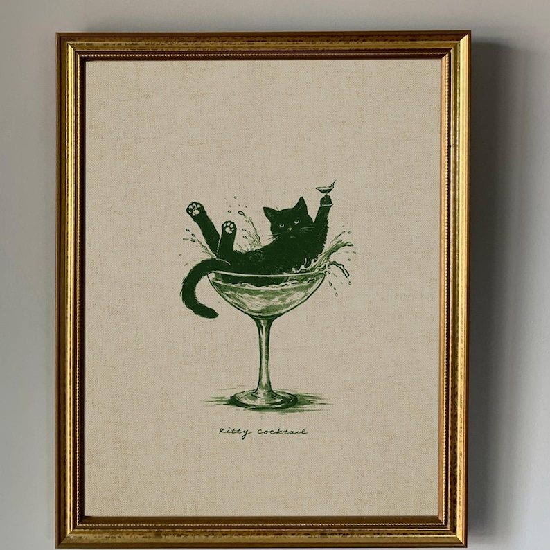 May include: Framed art print featuring a green illustration of a black cat lounging in a martini glass, with the words "Kitty Cocktail" below. The print is set against a neutral background and framed in a gold-colored frame.