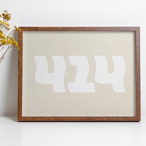 Peut inclure: Impression encadrée avec le chiffre "424" en blanc sur fond beige. Le cadre est en bois brun foncé. Un vase blanc avec des fleurs jaunes est à gauche du cadre.