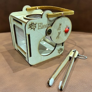 May include: A wooden bug box with a clear viewing window, a circular magnifying glass, and a small handle. The box has a hinged door with a ladybird design and the words "Bug Box". Includes wooden tweezers.