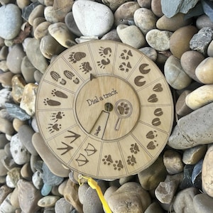 Peut inclure: Un cadran éducatif circulaire en bois clair portant l'inscription Dial a track avec un pointeur rotatif. La surface présente des illustrations gravées de diverses empreintes d'animaux, une icône de loupe et le mot Badger.